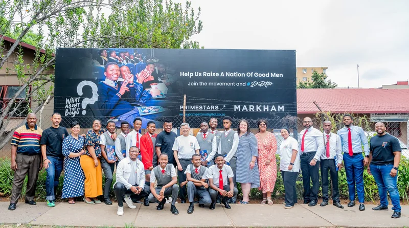 Learners and adults gathered in front of a What About The Boys and Markham programme billboard.