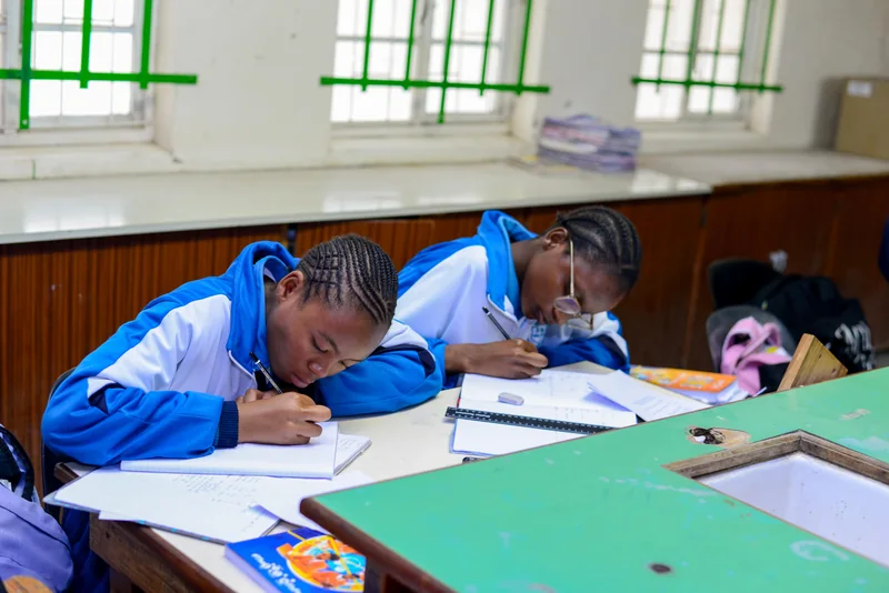 Two learners writing in notebooks at a classroom desk during EduCate at Ndengetho.