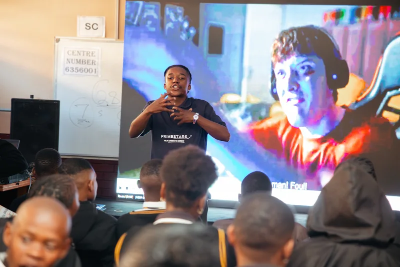 A facilitator wearing a Primestars shirt addressing seated learners beside a large screen.