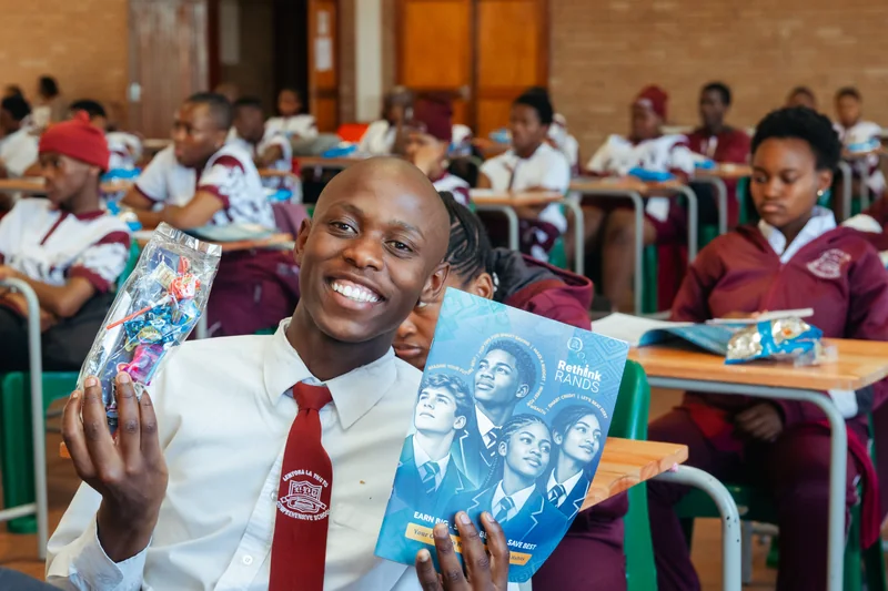A smiling learner holding a Rethink Rands booklet and a clear pack of sweets and pens, with classmates seated behind.