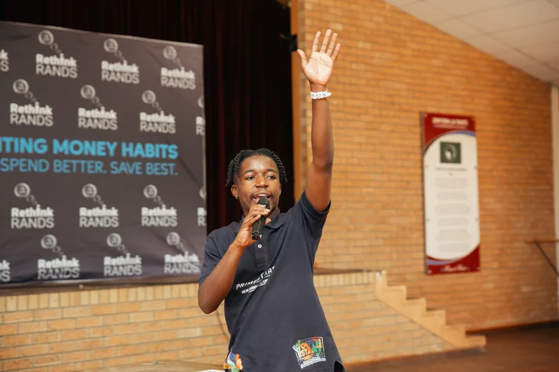 A presenter holding a microphone and raising one hand beside a Rethink Rands banner.