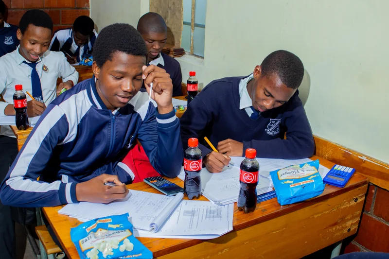 Learners writing on worksheets at desks with calculators, drinks and snack packets.