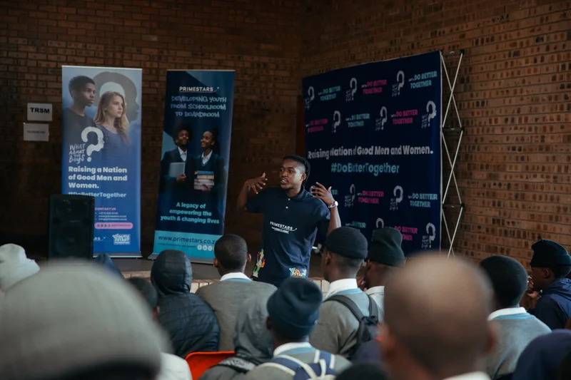 A facilitator speaking to learners in front of What About The Boys and Primestars banners.